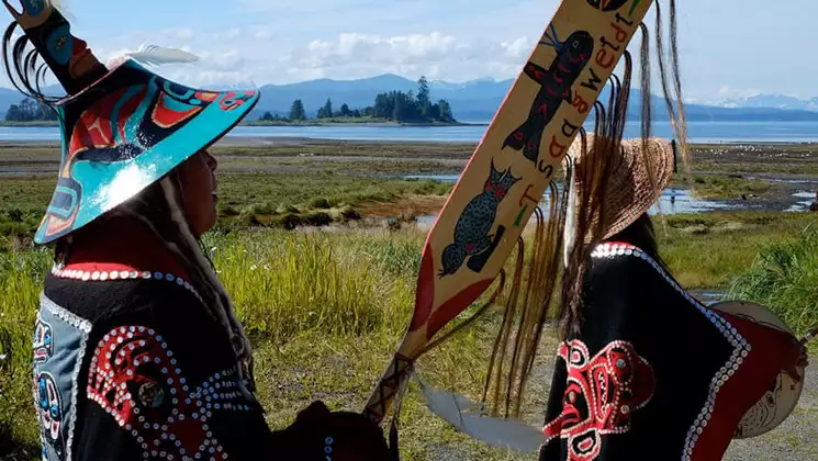 2 Native Alaskans stand on grassy shoreline wearing traditional Tlingit garb in turquoise, red & black with pearled buttons.