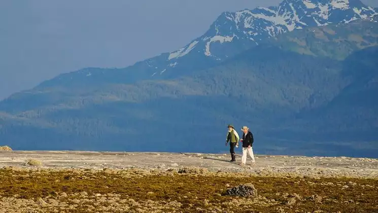 2 Wild Alaska Odyssey cruise travelers walk over tan & brown tundra backed by tall snowcapped peaks on a sunny day.