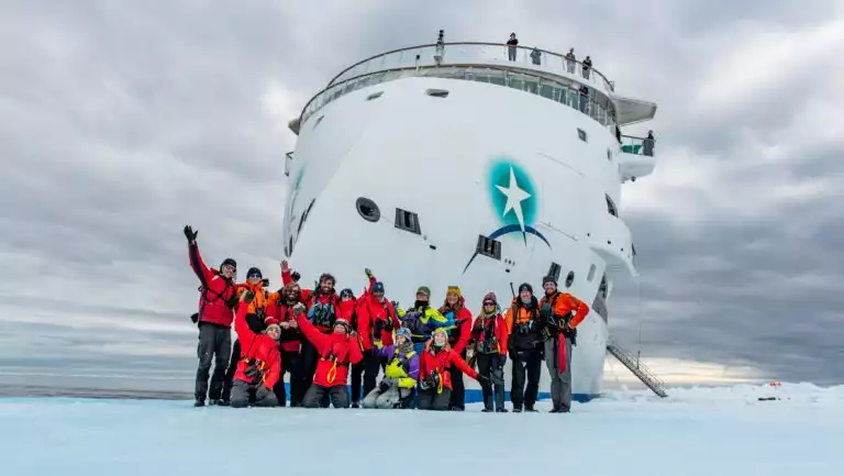 Group of expedition staff members in bright red jackets stand on fast ice in front of pointy bow of white expedition ship.