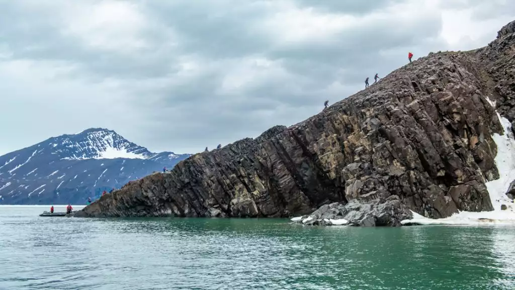 Small group of hikers walk up brown, columnar, rocky outcrop at water's edge as Zodiac boat sits below, in West Greenland.