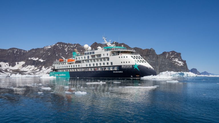Sylvia Earle small expedition ship with dark blue hull & white upper decks sits in calm sea with small iceberg bits floating by.