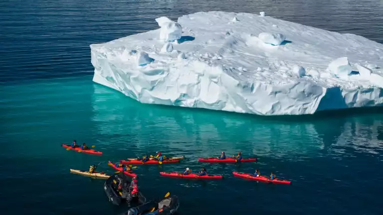 Kayakers in red and yellow kayaks stopped next to a large iceberg floating in the middle of the ocean