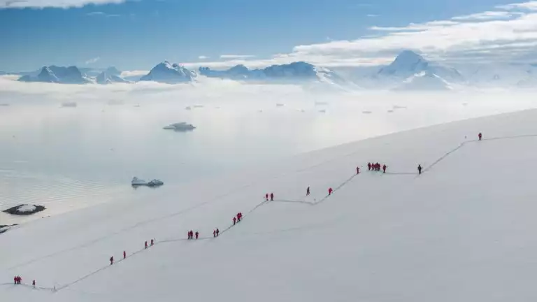 Travelers hike up a snowy hill in a zig zag position in front of breathtaking mountains surrounded by fog