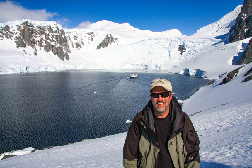 Man in open jacket, hat & sunglasses stands atop snowy mountainside with small expedition ship in calm bay below, in Antarctica.