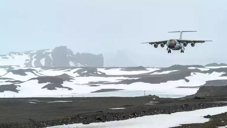 Small jet painted like a macaroni penguin comes in for a misty landing on a gravel airstrip among snowdrifts in Antarctica.