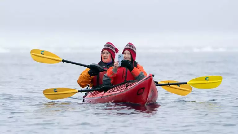 Woman & man tandem kayakers in red boat with yellow paddles pause to photograph the Antarctic icescape in calm water.