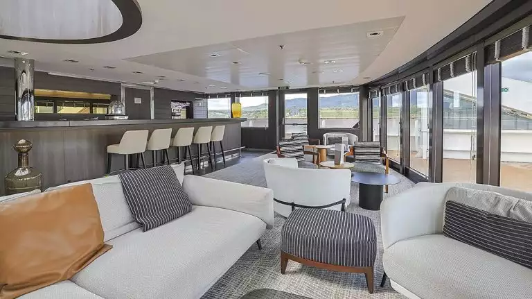 Living room aboard small ship cruise with tan couches and tan barstools at modern bar with large windows overlooking the ship
