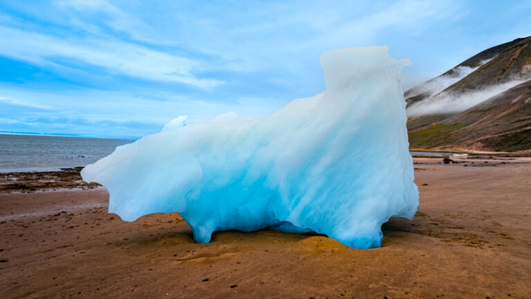 Bright blue iceberg bit washed up on brown shoreline under a blue sky in Baffin Bay, Greenland.