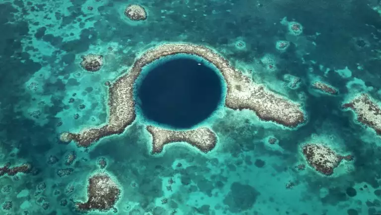 Aerial vie of Belize's Blue Hole dive site, a dark blue circle with tan coral reef & various shades of turquoise water around it.