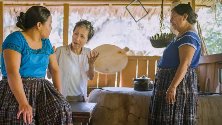 Female Belize & Honduras traveler talks with local women dressed in cultural garb in a primitive open-air kitchen.
