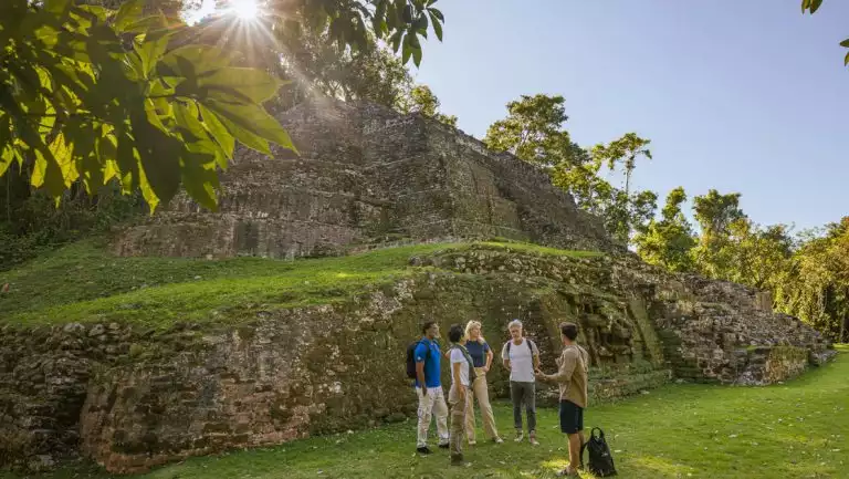 Small group of Belize & Honduras travelers stand with guide beside tall Maya ruins of stone & grass during a small ship cruise.