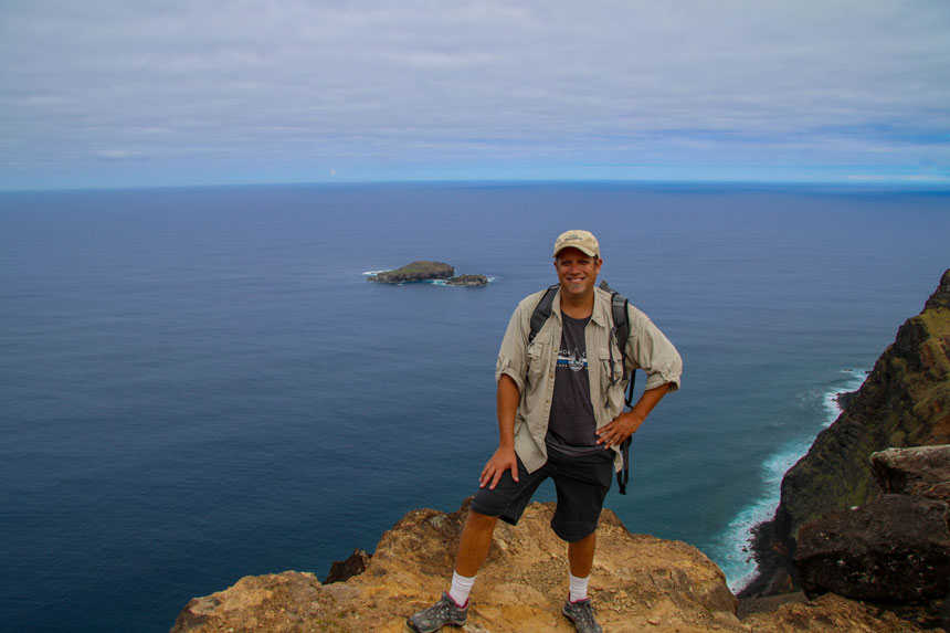 A traveler in a tan shirt and backpack stands on a cliff in Easter Island overlooking a small island in the ocean