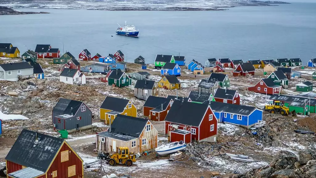 Brightly painted homes sit among tundra & snow on shoreline with small blue & white ship nearby in Ittoqqortoormiit, Greenland.