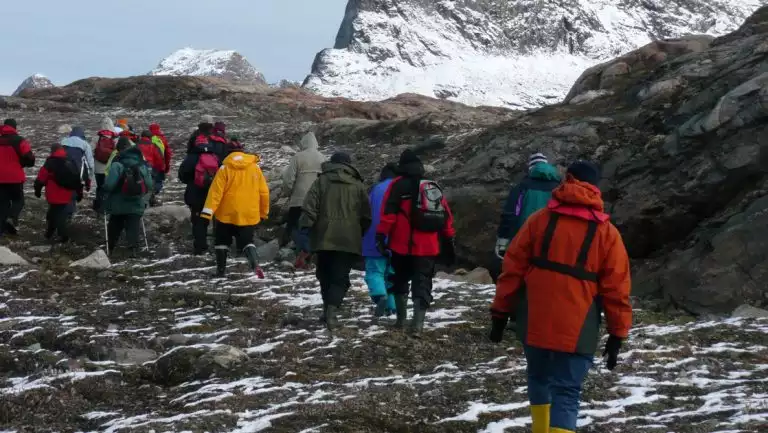 Line of east & south Greenland travelers in colorful jackets walks over tundra with snow patches under snow-covered peaks.