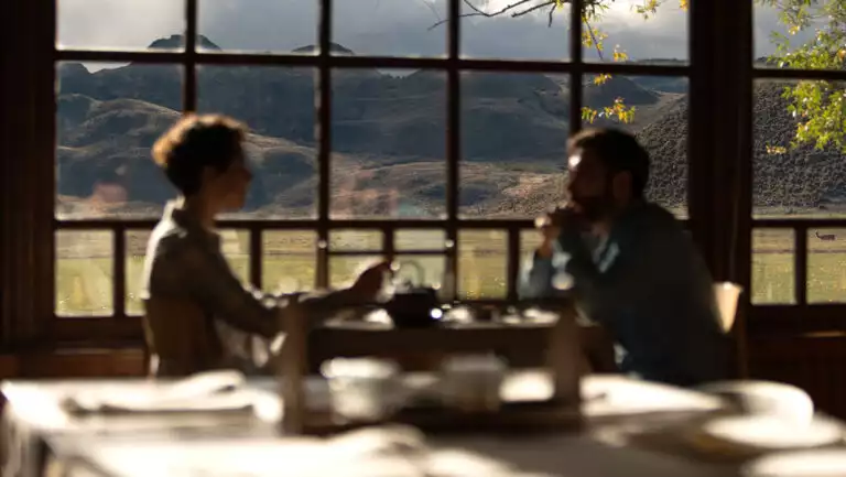 Man & woman dine at a 2-top table beside large window with wood lattice & views to green fields & mountains in Patagonia.