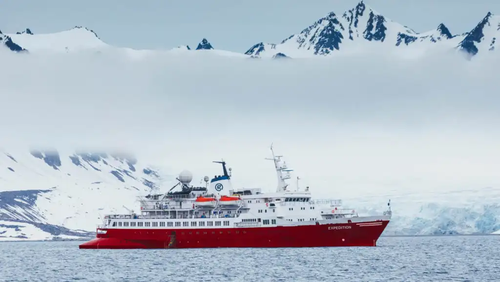 G Expedition ship with red hull & white upper decks sits in calm sea under clouds in snowy Antarctic landscape.