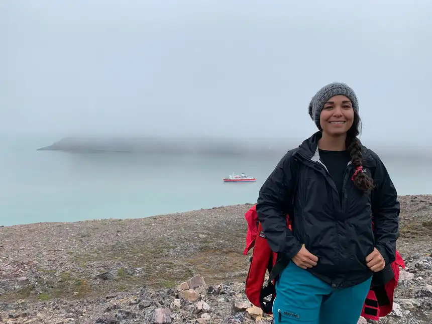 Woman in black jacket & blue waterproof pants stands atop rocky mountain's edge with small red & white MS Expedition ship behind in the fog.
