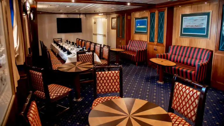 G Expedition ship club bar with wood tables, red & white chairs, striped couches, blue carpet & view windows.