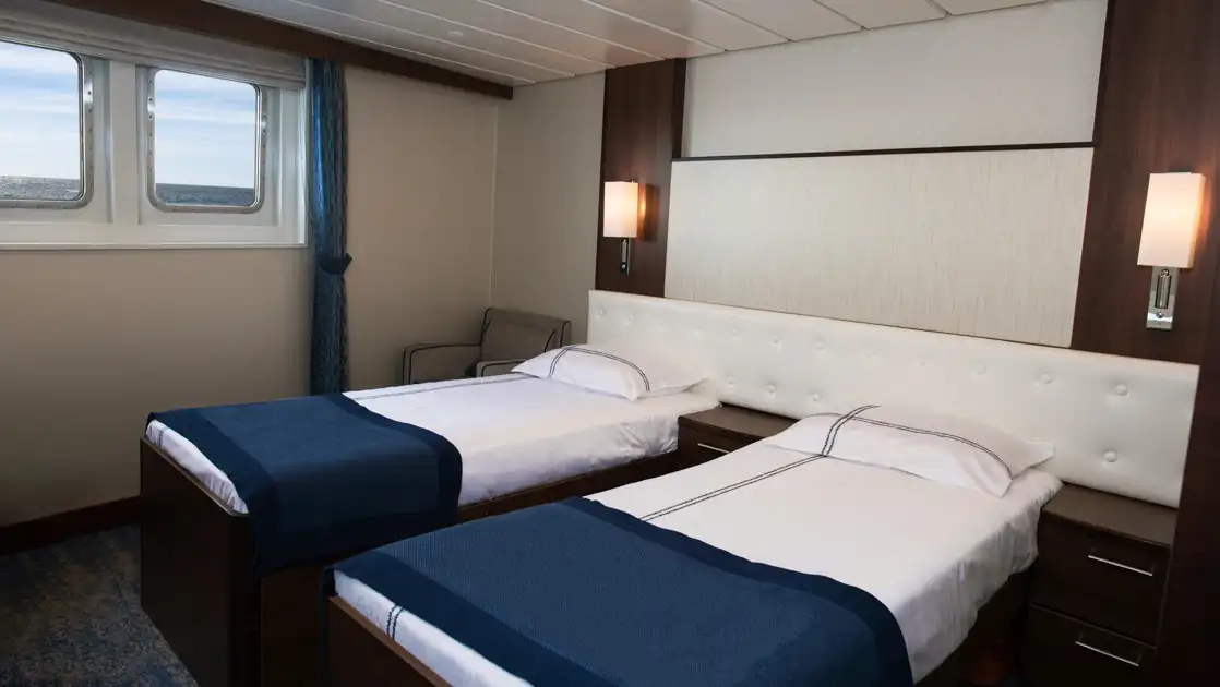 Deluxe Twin cabin on G Expedition ship with 2 twin beds in blue & white linens, padded white headboard & 2 small windows.