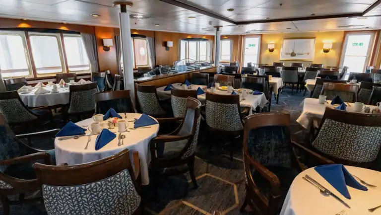 Dining room of G Expedition ship, with 4- & 6-person round tables set with white & blue linens & silver cutlery.
