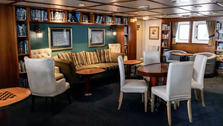 Library with white, padded chairs, filled wooden bookshelves & wooden tables aboard G Expedition ship.