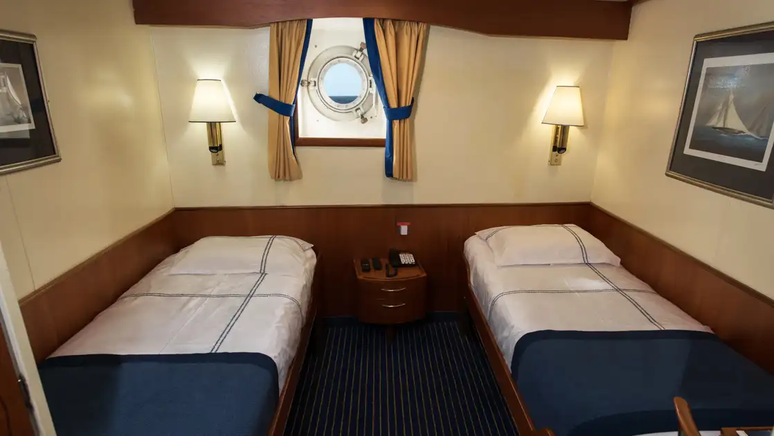 Lower Deck Twin cabin on G Expedition ship with 2 twin beds in blue & white linens, wood accents & 1 porthole.