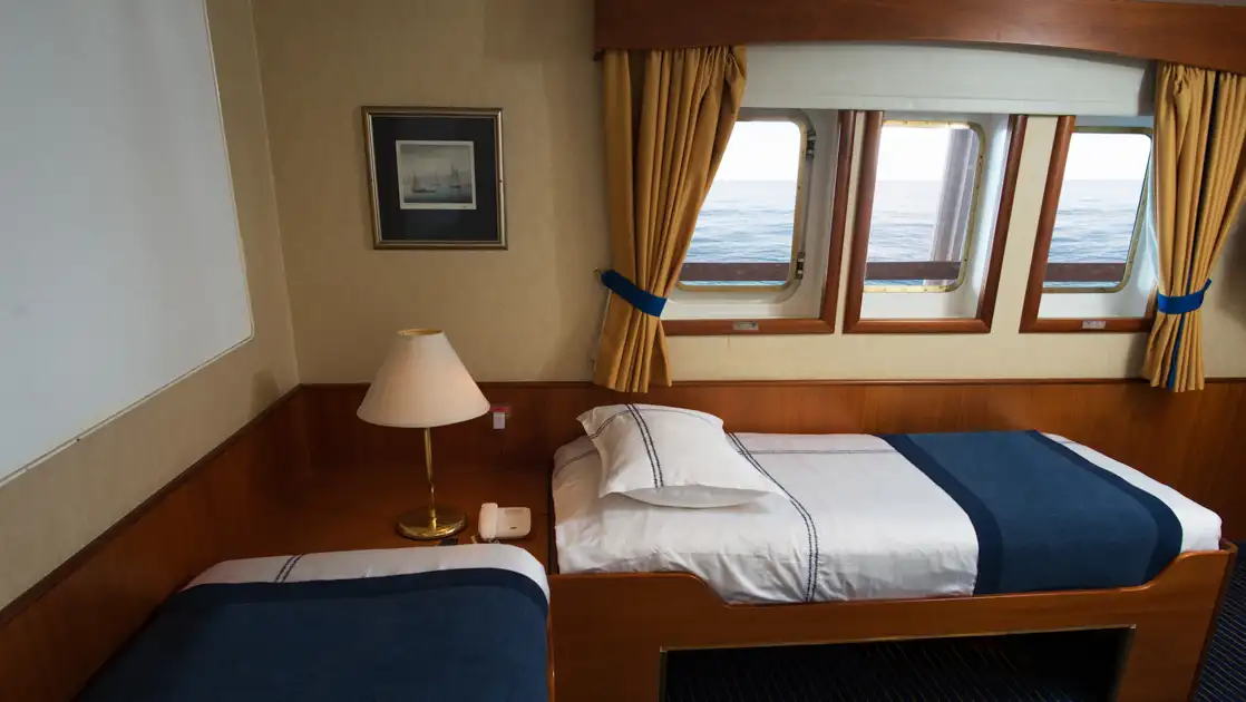 Superior Twin cabin on G Expedition ship with 2 L-shaped twin beds in blue & white linens, wood accents & 3 small windows.