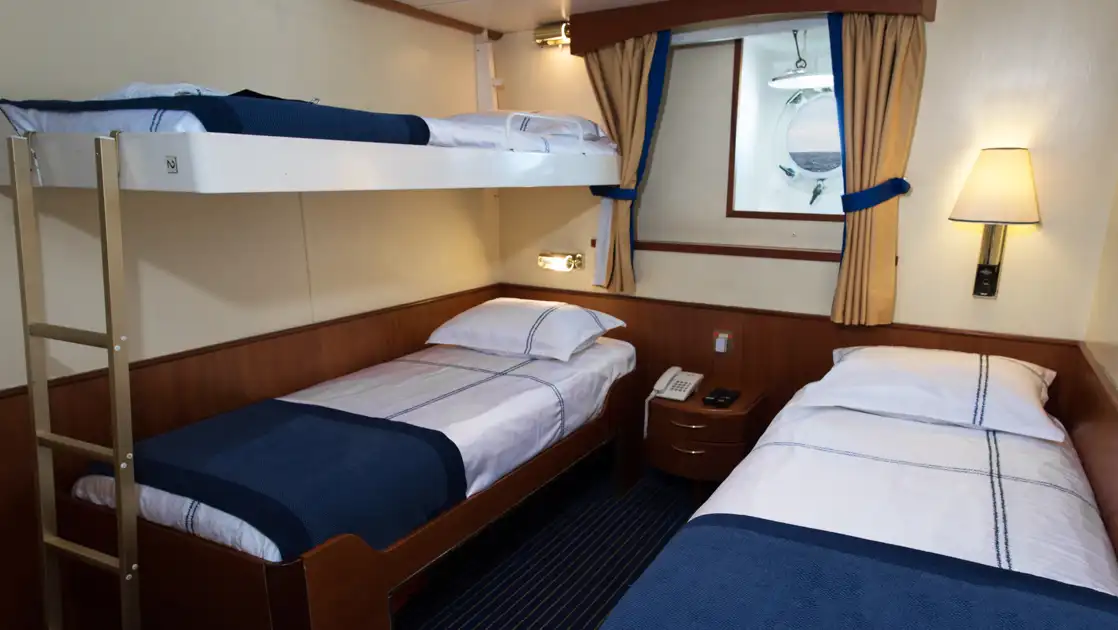 Main Deck Triple cabin on G Expedition ship with bunkbeds & 1 lower twin bed in blue & white linens, wood accents & porthole.