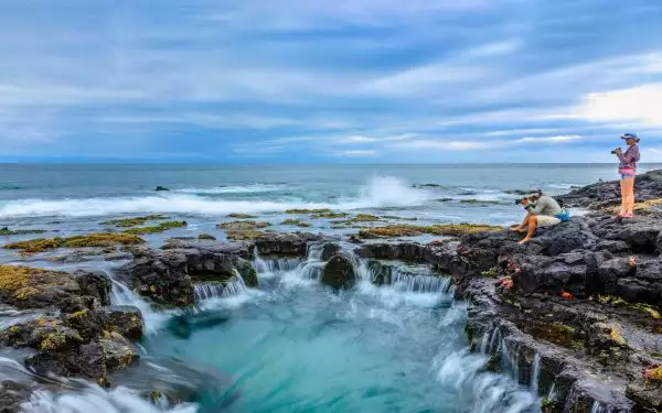 A little girl in a hat and purple jacket holds a camera while standing on lava rock by a seated adult taking photographs of the ocean and a tidepool