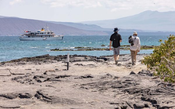 Two travelers hike on lava rock on the shore in the Galapagos Islands with a private yacht at sea just off shore