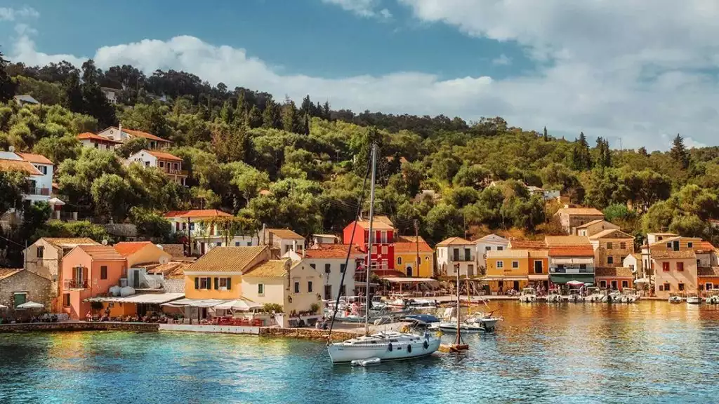 Yellow, orange and red houses and buildings dot the harbor front in Gaios, Ithaca, with a sailboat docked in front of them.