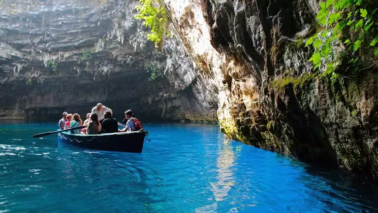 Travelers in a small row boat with a man in a white shirt rowing seen in a cave at Mellisani Lake in Greece with foilage growing on the steep walls