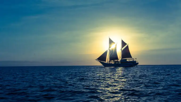 Indonesian sailing ship with sails unfurled cruises while silhouetted by a sunbeam on a calm day.