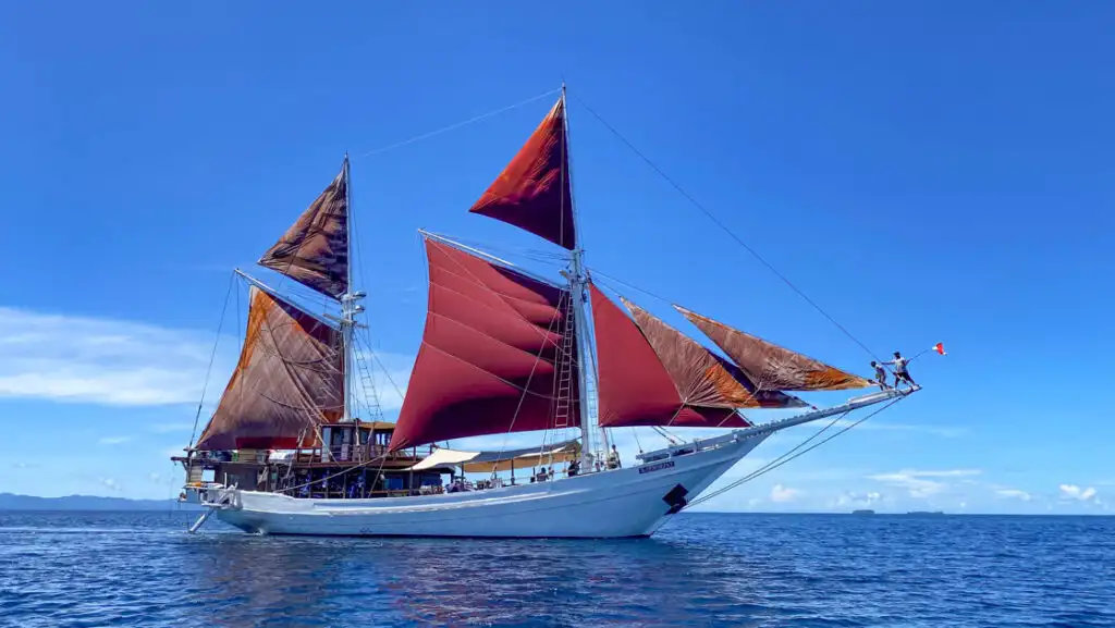 Katharina Indonesia sailing ship with wooden hull painted white & large burgundy sails unfurled on a sunny day at sea.
