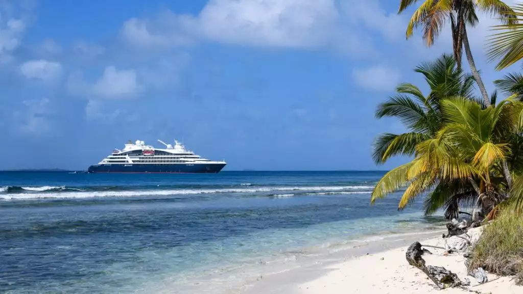 Small modern expedition ship with blue hull & white upper decks sits offshore from white-sand beach with palm trees in Panama.
