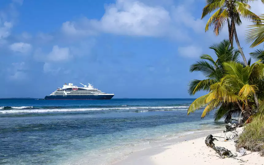 Small modern expedition ship with blue hull & white upper decks sits offshore from white-sand beach with palm trees in Panama.