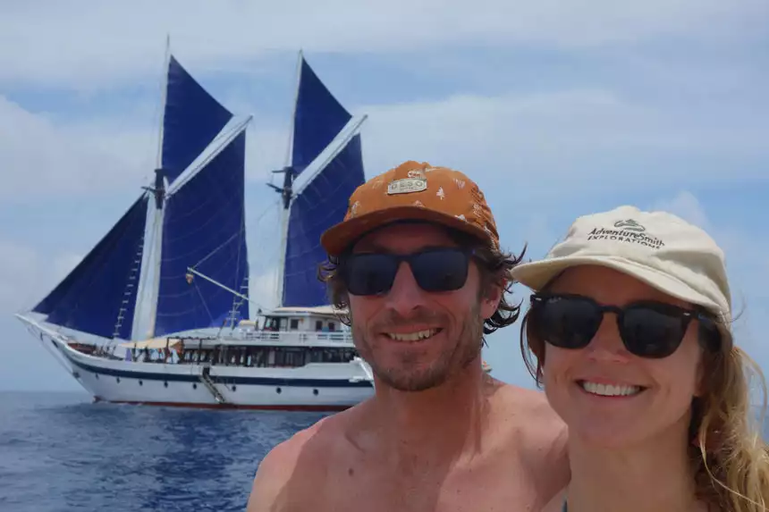 Man & woman in baseball caps & sunglasses pose for a photo with the Ombak Putih Indonesian sailing ship behind in white with blue sails.