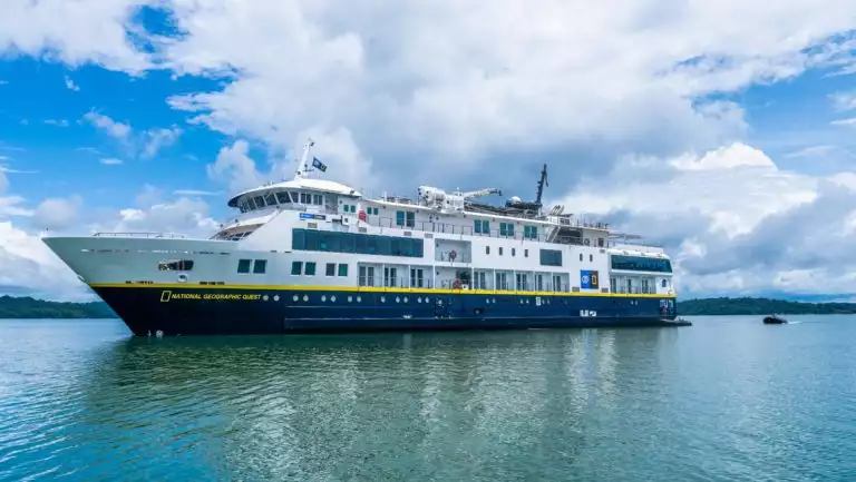 National Geographic Quest small ship in blue hull & white upper decks sits in calm sea under sun at Barro Colorado, Panama.