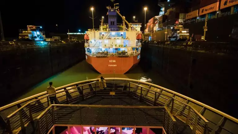 Guest stands on bow of small ship Nat Geo Quest as she transits the Panama Canal behind a cargo ship with red hull at night.