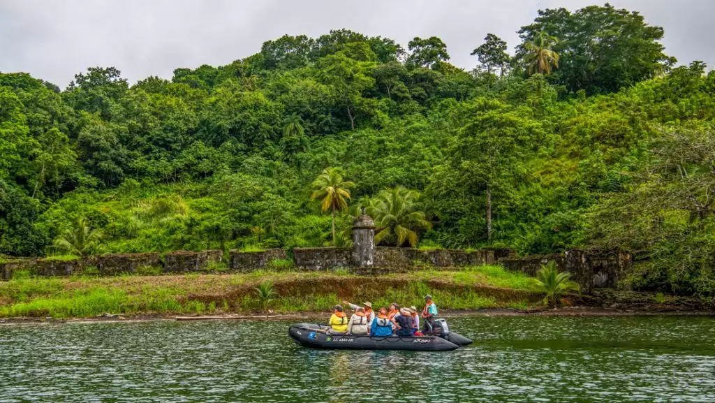 Zodiac boat with Panama canal travelers motors past aging historic fort walls backed by jungle as a guide points.