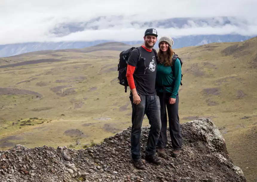 Man & woman stand atop a rocky overlook on a hike in Patagonia with green tundra below & snowy peaks behind.