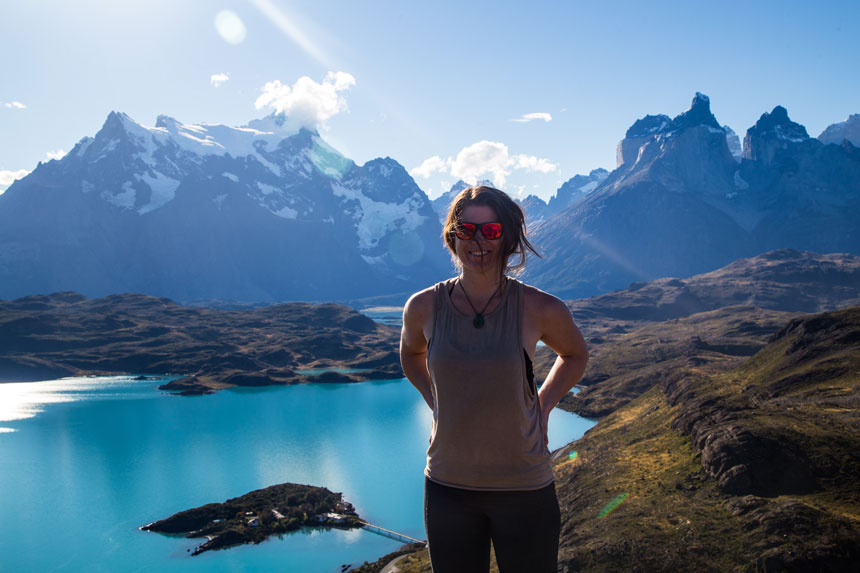 Woman in gray tank top & reflective red sunglasses stands atop grassy peak overlooking teal lake below & jagged snowy peaks behind, in Patagonia.