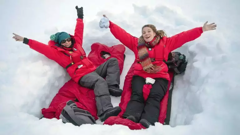 Two travelers pose for a picture in their snow camping set up with red sleeping bags, red jackets and adventure clothing.