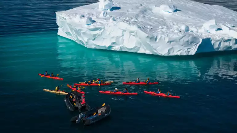 Aerial view of yellow & red kayaks & black Zodiacs in teal calm sea beside a snowy iceberg bit on a sunny day in Antarctica.