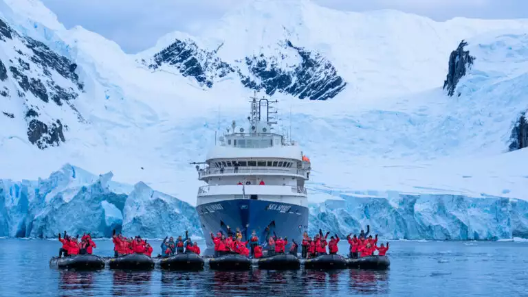 Line of Zodiac boats with Antarctica travelers in red jackets sits in front of Sea Spirit small ship & snowy mountains behind.