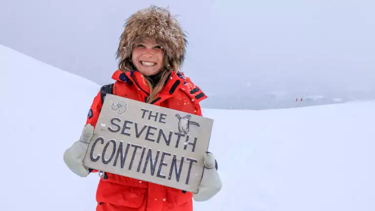 Woman in red parka, white mitts & fake fur hat holds sign reading The Seventh Continent atop a snowfield in an Antarctic storm.