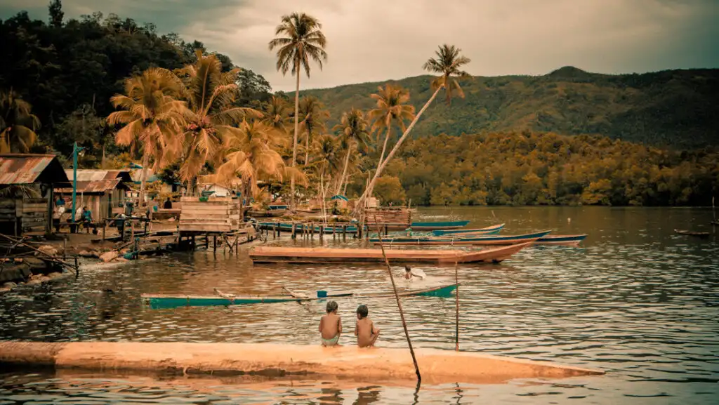 2 boys sit on floating dock overlooking small Indonesian seaside village & harbor under palm trees & green mountains.