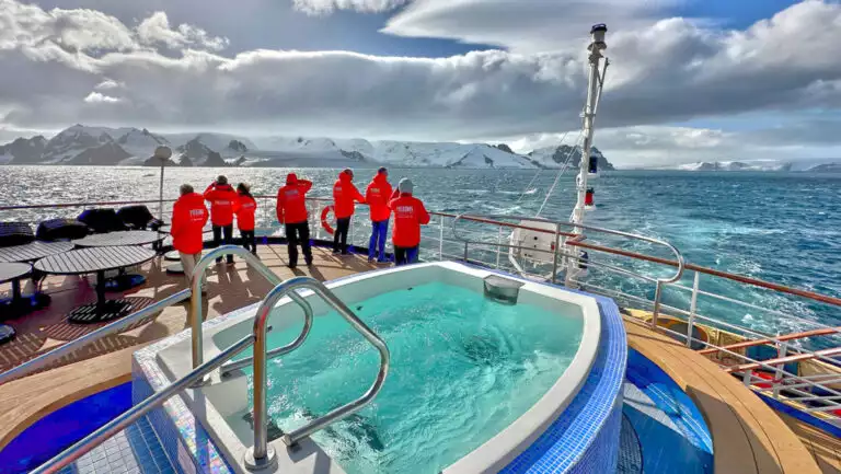 Small group of polar travelers in red jackets stand on deck of Sea Spirit small ship, beside large Jacuzzi in blue tile.