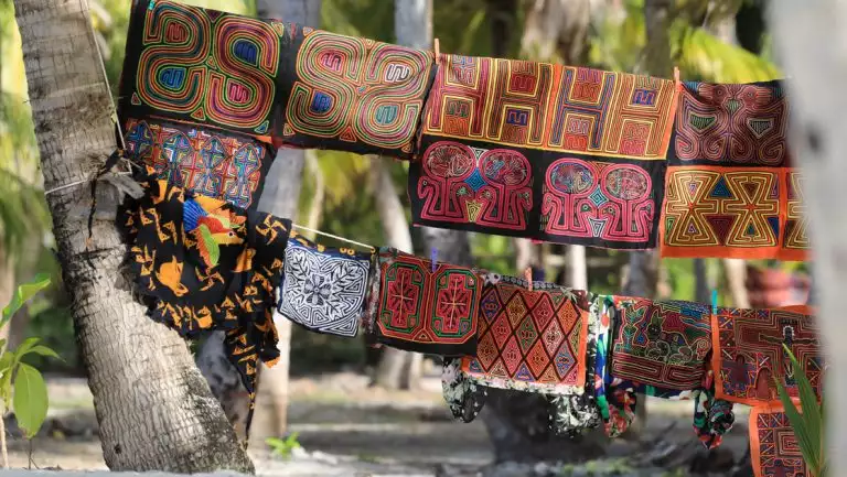 Multicolored, bright, woven textile squares hang from rope between palm trees over white-sand beach in Panama.