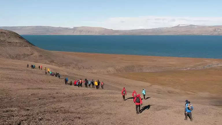 Line of South Spitsbergen cruise guests in various colorful coats hike over arid tundra near the sea on a sunny day.